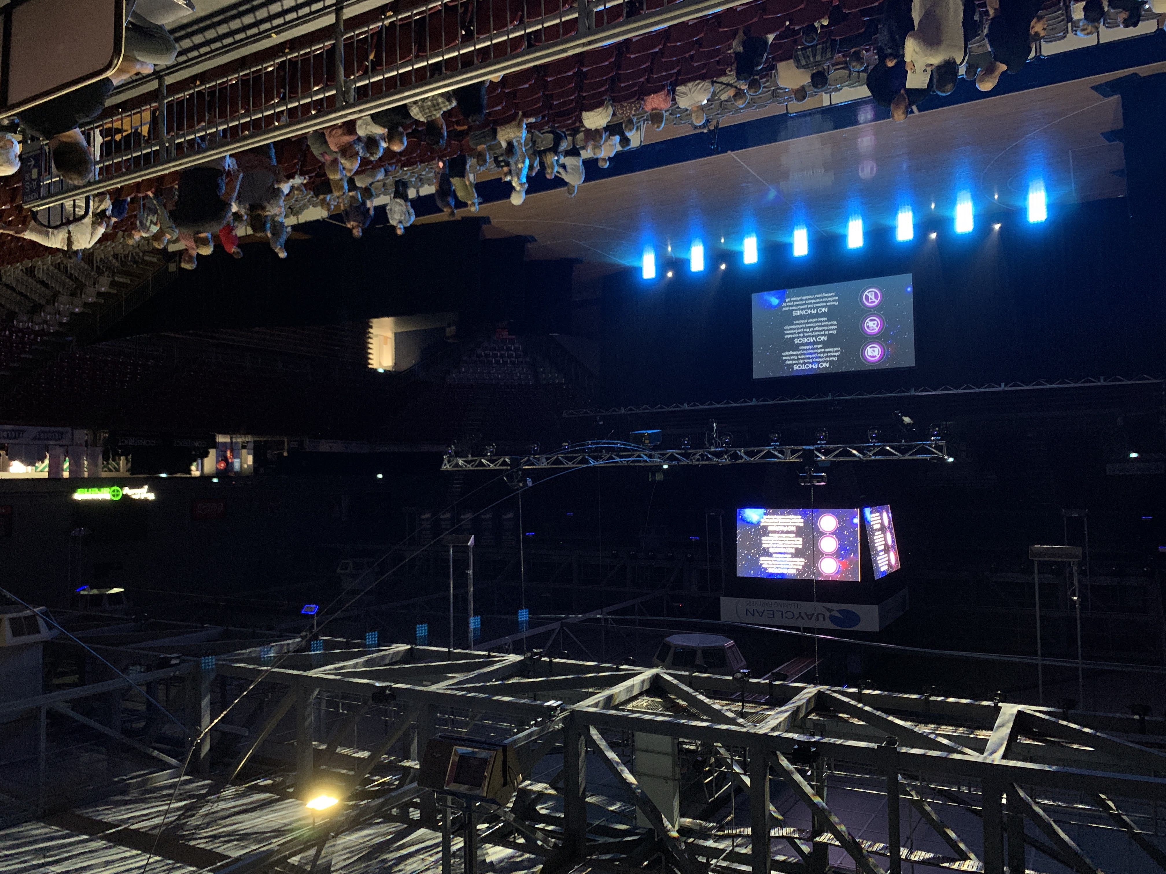 This image depicts an indoor arena setting during what appears to be the preparation for an event. The venue is semi-lit with a combination of natural and artificial lighting. In the upper part of the image, we can see a complex network of metal trusses, some with attached stage lights, that form part of the ceiling structure. There is a central, cubic scoreboard displaying information, which indicates that the venue can be used for sports events as well.

Below, a large stage is visible with a screen that reads "NO PHOTOS, NO VIDEOS, NO PHONES". This message suggests that there may be a performance or presentation planned that requires undivided attention or has restrictions on recording. The blue stage lights are on, providing an accent to the stage area and drawing focus. The seating area, which consists of rows of fixed red seats, is mostly empty, with just a few individuals gathered on the floor and around the stage, likely event staff or early attendees. The presence of people in casual attire indicates an informal gathering at this time, perhaps before the main event commences.

The layout and current activity suggest the image was taken during the preparation or soundcheck before an event. This setup would be particularly relevant for someone involved in event management or audiovisual equipment installation.