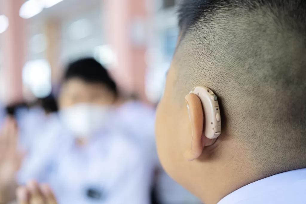 Close-up of a person with a hearing aid, focusing on the device behind their ear, with a blurred background showing another person in a white mask and uniform, participating in an event. 