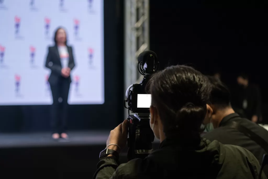  A videographer capturing a speaker on stage during a live event, demonstrating the behind-the-scenes work involved in professional live streaming. The focused camera and blurred background highlight the speaker, showcasing the technical expertise required to deliver high-quality live streaming services.